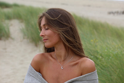 Une femme aux longs cheveux bruns se tient debout sur une plage de sable, portant le collier Ayla | Pendentif Lait Maternel Or de Mama Jewels avec son haut gris à épaules dénudées. Elle regarde sur le côté avec les yeux fermés, encadrée par de hautes herbes et un océan flou en arrière-plan.