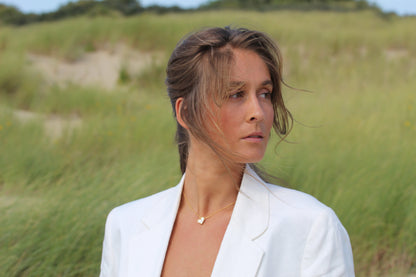 Une femme aux cheveux châtain clair portant un blazer blanc et le collier Louisa Lait Maternel Or de Mama Jewels se tient dans un champ herbeux, regardant sur le côté avec une expression sereine sur fond de dunes de sable et de verdure.